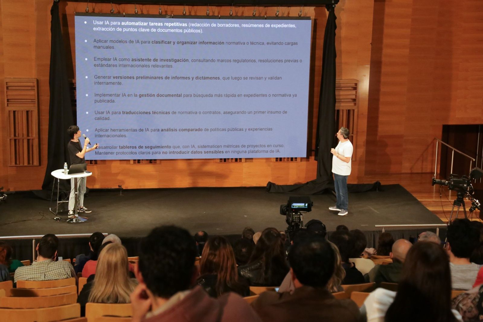 Foto de un escenario con una pantalla grande proyectada. La lamina enumera usos concretos de inteligencia artificial en el Estado: automatizar tareas repetitivas como redaccion de borradores y resumenes de expedientes, clasificar y organizar informacion normativa, usarla como asistente de investigacion sobre marcos regulatorios, generar versiones preliminares de informes y dictamenes, gestion documental, traducciones tecnicas de normativa, analisis comparado de politicas publicas y tableros de seguimiento. El ultimo punto advierte que hay que mantener protocolos claros para no introducir datos sensibles en ninguna plataforma. A los costados del escenario, dos personas con microfono. El publico mira desde las butacas.