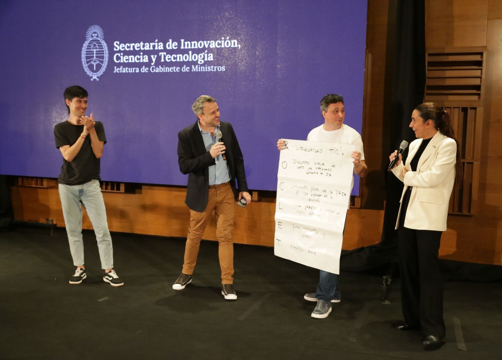 Foto del cierre de la jornada sobre el escenario. Cuatro personas de pie: una aplaude, dos tienen microfono y una sostiene con las dos manos un papelografo grande escrito a mano con marcador. En el margen izquierdo bajan las letras R, O, C, L, E y T, y al lado de cada una hay una linea anotada: es la estructura del discurso de cierre, armada con el metodo ROCLET. Detras, sobre fondo violeta, el escudo nacional y el texto Secretaria de Innovacion, Ciencia y Tecnologia, Jefatura de Gabinete de Ministros.
