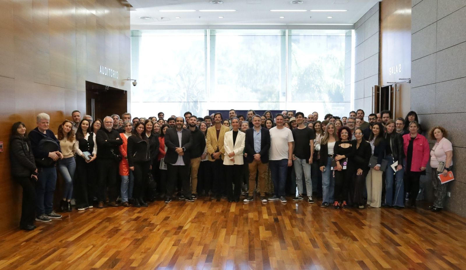 Foto grupal en el hall del edificio, delante de un ventanal. Alrededor de cien personas de pie, en varias filas, mirando a camara. Algunas sostienen cuadernos y carpetas. A los costados se ven las puertas del auditorio y de la sala 2.