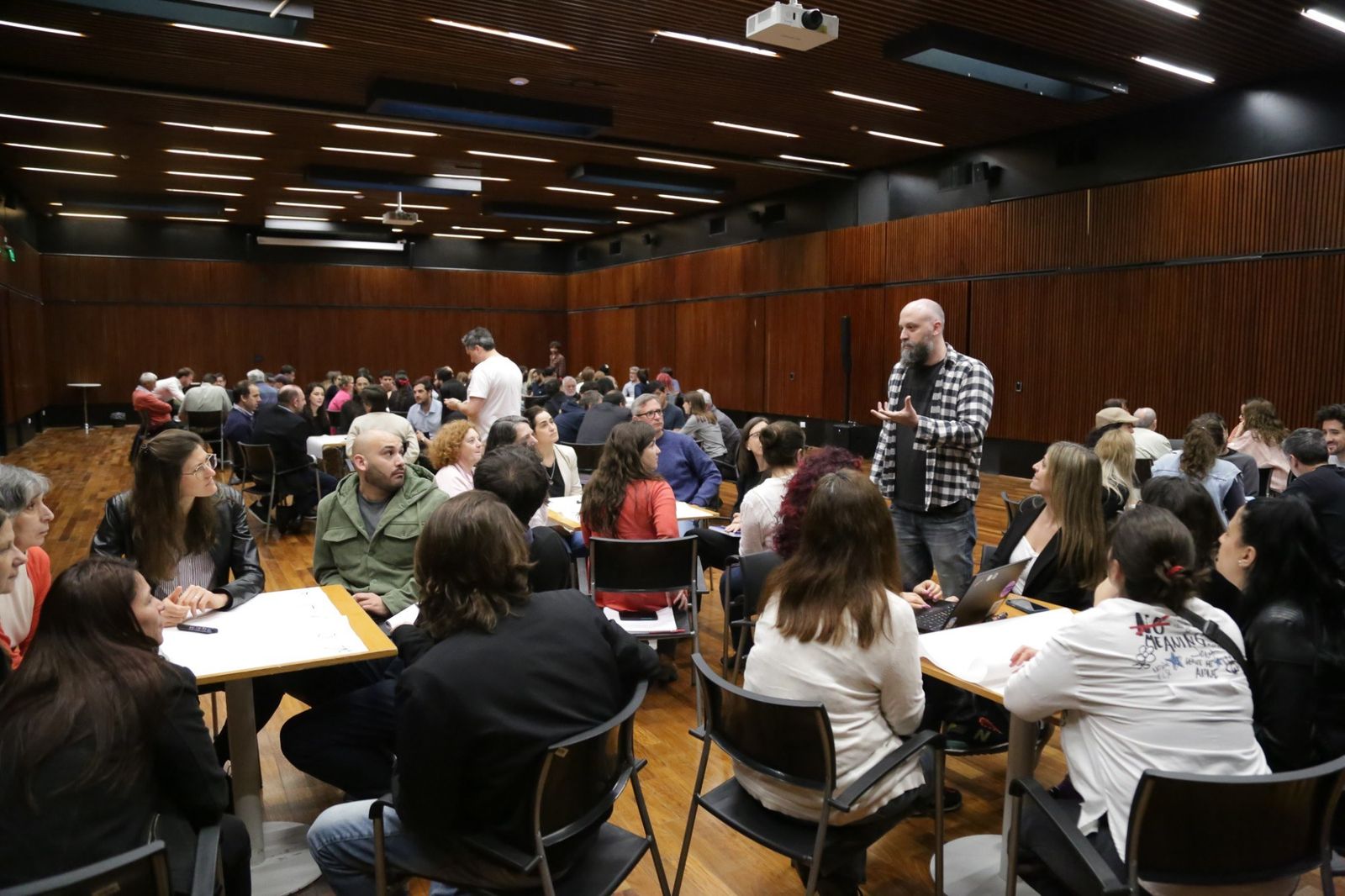 Foto de un salon con mesas de trabajo. Alrededor de cada mesa hay grupos de personas sentadas, con hojas grandes de papel, cuadernos y notebooks abiertas. Un hombre de camisa a cuadros esta de pie junto a una de las mesas, explicando algo con las manos, y las personas sentadas lo miran. Al fondo se ven mas mesas ocupadas. Nadie mira hacia adelante: todos trabajan hacia adentro de su grupo.